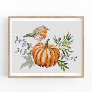 May include: A cross-stitch pattern depicting a small bird perched atop an orange pumpkin. The pumpkin is surrounded by green leaves and purple flowers.