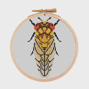 May include: A cross-stitch embroidery of a cicada insect centered in a round wooden hoop. The cicada features a yellow and tan body with black wing details and prominent red eyes, stitched onto a white fabric background with a visible grid pattern.