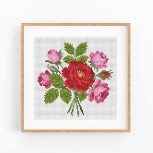 May include: A square framed cross-stitch embroidery featuring a central bouquet of flowers. The design displays one large red rose surrounded by several smaller pink roses and rosebuds, all accented with green leaves and stems on a white grid.