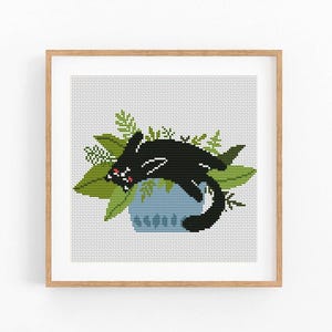 May include: A framed cross-stitch art piece featuring a black cat lounging on a blue pot surrounded by green leaves. The cat has red accents in its mouth and eyes. The artwork is set against a light gray background.