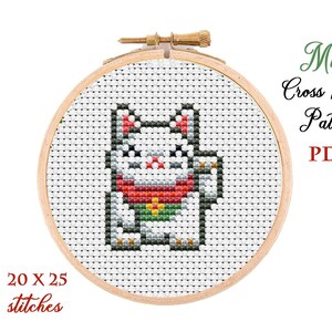 May include: A white cat with red and green clothing, holding a gold coin, is depicted in a cross stitch pattern on a wooden embroidery hoop. The pattern is 20 x 25 stitches. The text "Mini Cross Stitch Pattern PDF" is written in green and red.