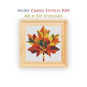 Puede incluir: Un patrón de punto de cruz con una hoja de arce multicolor en tonos rojos, naranjas, amarillos y verdes sobre una cuadrícula blanca. El texto dice MINI CROSS STITCH PDF 48 x 50 STITCHES. El diseño se muestra en un marco de madera clara.