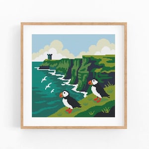 May include: Framed pixel art print of a coastal scene with two puffins. The image shows green cliffs, a turquoise sea, and a blue sky with clouds. The puffins have black and white bodies, orange beaks, and red feet.