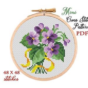 May include: A cross-stitch pattern of a bouquet of purple violets with green leaves and a yellow ribbon, set in a wooden embroidery hoop. The text "Mini Cross Stitch Pattern PDF" and "48 x 48 stitches" are also visible.