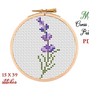Può includere: Uno schema di punto croce per un fiore viola con foglie verdi, mostrato in un telaio da ricamo in legno. Lo schema è largo 15 punti e alto 39 punti. Il testo "Mini Cross Stitch Pattern PDF" è visualizzato sopra il telaio.