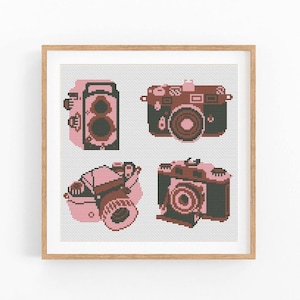 May include: A square cross-stitch pattern featuring four distinct camera designs rendered in shades of pink, dark brown, and black. The cameras are arranged in a grid layout against a white grid background, all enclosed within a light wood frame.