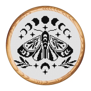 Puede incluir: Diseño de bordado en blanco y negro de una polilla con las alas extendidas, rodeada de lunas crecientes y estrellas. El diseño está enmarcado en un aro de madera.