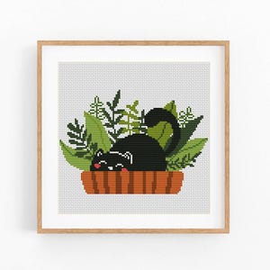 May include: Cross-stitch art of a black cat resting in a brown planter filled with green leaves. The cat has a white stripe and pink cheeks. The planter has vertical orange stripes. The artwork is framed in a light brown wooden frame.