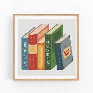 May include: A framed cross-stitch artwork featuring a stack of colorful books. The books are in shades of blue, red, yellow, and green, with the word "ENCYCLOPEDIA" visible on one spine and "NOVELS" on another. The artwork is set against a white background.