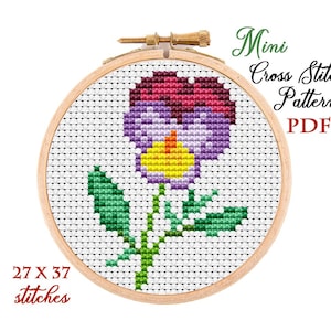 May include: A cross-stitch pattern of a pansy flower in a wooden embroidery hoop. The flower is purple, yellow, and maroon, with green leaves and stem. The text "Mini Cross Stitch Pattern PDF" is in green script. The pattern is 27 x 37 stitches.