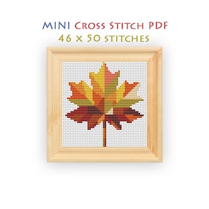 May include: A mini cross stitch PDF pattern featuring a maple leaf design with 46 x 50 stitches. The leaf is rendered in a gradient of yellow, orange, red, and green tones, displayed within a light-colored wooden square frame on a white background.