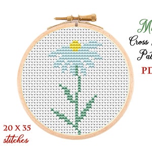 May include: A cross stitch pattern for a blue flower with a yellow center and green leaves. The pattern is 20 stitches wide by 35 stitches tall. The pattern is displayed in a wooden embroidery hoop.