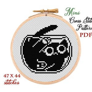 May include: A cross-stitch pattern in a wooden hoop features a black cat design with glasses. The text "Mini Cross Stitch Pattern PDF" is displayed, along with "47 X 44 stitches". The design is on white fabric.