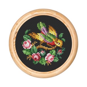 May include: A framed cross-stitch embroidery depicting a colorful bird perched on a branch surrounded by pink and green flowers.