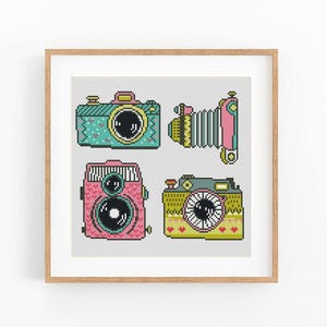 May include: A cross-stitch pattern featuring four distinct cameras in a light wooden frame. The cameras are rendered in pixelated style with colors including teal, pink, yellow, and grey, showcasing various lens designs and body shapes.