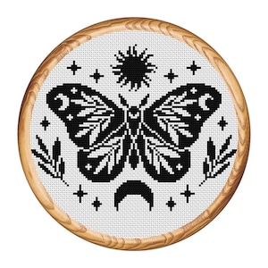 May include: A black and white cross-stitch design featuring a moth with celestial elements, including a sun, stars, and a crescent moon. The design is set within a round wooden frame, creating a rustic and handcrafted aesthetic.