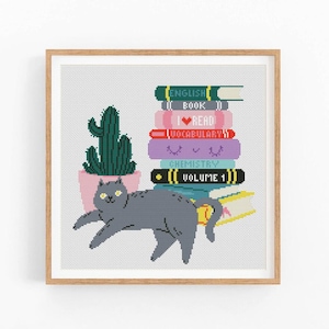 Puede incluir: Un diseño de punto de cruz con un gato gris tumbado cerca de una pila de libros y un cactus en maceta. Los libros están etiquetados como "English Book", "I Read", "Vocabulary", "Chemistry" y "Volume 1". El gato tiene ojos amarillos.