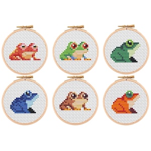 Könnte beinhalten: Sechs kleine Kreuzstich-Stickrahmen mit jeweils einem anderen bunten Frosch-Design. Die Frösche sind in Rot-, Grün-, Blau- und Brauntönen gehalten, mit Details wie roten Augen und kontrastierenden Bäuchen. Die Rahmen sind aus Holz.