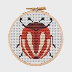 May include: A circular wooden embroidery hoop containing a cross-stitch pattern of a beetle. The beetle features a dark brown head, a vibrant red body with symmetrical cream-coloured wing markings, and thin black legs against a white fabric.