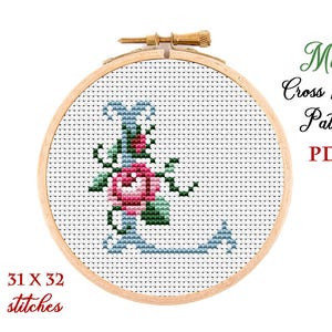 May include: A cross-stitch pattern in a wooden hoop features the letter "L" decorated with a pink rose and green leaves. The design includes blue accents and is labeled "Mini Cross Stitch Pattern PDF". The pattern is 31 x 32 stitches.