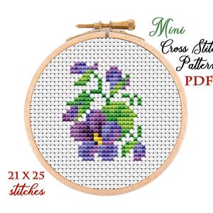 May include: A cross-stitch pattern of a floral design in a wooden hoop. The design features purple and green flowers with a yellow center. The text "Mini Cross Stitch Pattern PDF" and "21 x 25 stitches" are also visible.