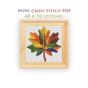 May include: A cross stitch pattern of a maple leaf in a light wood frame. The leaf features a gradient of yellow, orange, red, green, and blue colors. The text above reads MINI CROSS STITCH PDF 48 x 50 STITCHES. The design is on a white grid.
