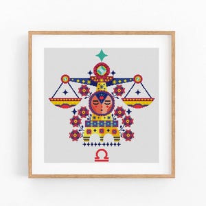 May include: A framed cross-stitch artwork featuring the Libra zodiac symbol. The design includes a face, scales, and floral elements in red, yellow, blue, and green. The artwork is set against a light gray background and framed in a light brown wooden frame.