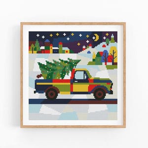 May include: A cross-stitch pattern in a light wood frame depicting a multicoloured pickup truck carrying a green Christmas tree. The background features a snowy landscape with colourful houses, trees, and a dark blue night sky with a yellow crescent moon.