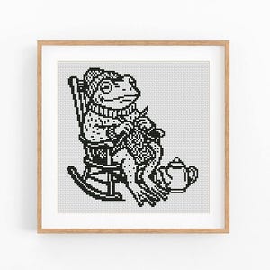 May include: A black and white cross-stitch pattern featuring a frog wearing a knit hat and sweater, sitting in a rocking chair while knitting with needles. A small teapot sits on the floor next to the chair, all framed in a light wood frame.
