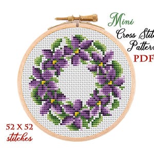 May include: A cross-stitch pattern of a violet flower wreath in a wooden embroidery hoop. The design features purple flowers with green leaves. Text reads "Mini Cross Stitch Pattern PDF" and "52 x 52 stitches".