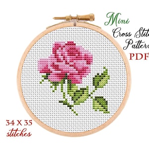 May include: A cross-stitch pattern of a pink rose in a wooden embroidery hoop. The design includes green leaves and the text "Mini Cross Stitch Pattern PDF". The pattern is 34 x 35 stitches.