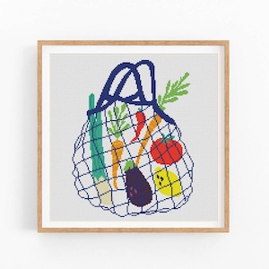 May include: A framed cross-stitch artwork depicts a blue mesh shopping bag filled with colorful vegetables. The bag contains carrots, a tomato, a lemon, an eggplant, and other produce. The artwork is set against a white background.