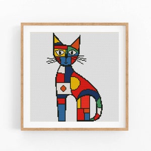 May include: A framed cross-stitch art piece featuring a colorful, geometric cat design. The cat is composed of various shapes and colors, including red, blue, yellow, and green. The artwork is set against a white background and framed in a light brown wooden frame.