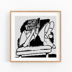 May include: A black and white illustration of a person reading a book, with the text "READ MORE BOOKS" on the cover. The person is reclining on a couch. The artwork is framed in a light brown wooden frame.