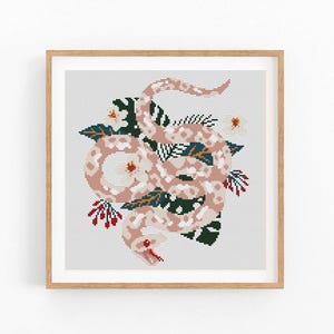 May include: A cross-stitch pattern featuring a pink snake with floral details. The snake is curled around a cluster of white flowers and green leaves. The background is a light gray.