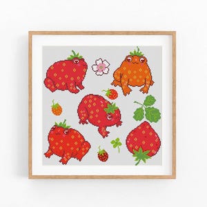 May include: A square cross-stitch pattern in a light wood frame featuring three frogs with red and orange strawberry-patterned skin, a single strawberry, a small white flower, green leaves, and a clover, all set against a light gray grid background.