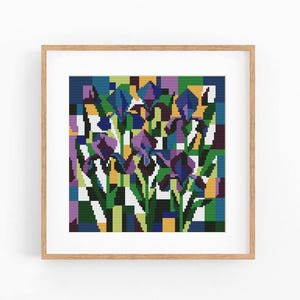 May include: A square framed cross-stitch pattern featuring a pixelated floral design of purple and blue irises with green stems and leaves, set against a geometric background of yellow, blue, and white blocks in a light wood frame.