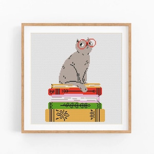 Puede incluir: Ilustración de punto de cruz de un gato gris con gafas rojas, posado sobre una pila de libros coloridos. Los libros son rojos, rosas, verdes y amarillos. La obra está enmarcada en un marco de madera marrón claro.