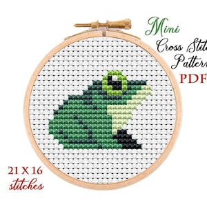 Könnte beinhalten: Ein Kreuzstichmuster einer grünen Frosch in einem hölzernen Stickrahmen. Der Text "Mini Cross Stitch Pattern PDF" und "21 x 16 Stiche" sind ebenfalls enthalten. Der Frosch besteht aus verschiedenen Grüntönen.