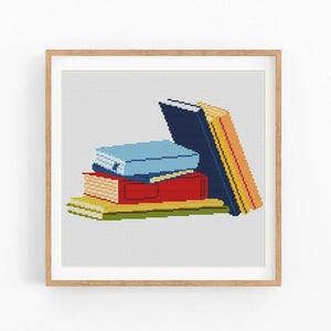 May include: A framed cross-stitch artwork featuring a stack of books. The books are in various colors, including blue, red, yellow, and orange. The artwork is set against a white background and framed in a light brown wooden frame.