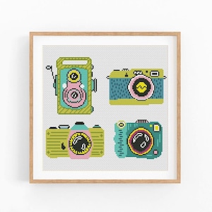 May include: A cross-stitch pattern featuring four vintage-style cameras in a grid layout. The cameras are rendered in pixel art style with vibrant colors including lime green, teal, pink, and yellow, all framed within a light wood square frame.