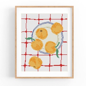 May include: A framed cross-stitch artwork featuring a still life of oranges on a plate. The plate has a blue rim and sits on a white and red checkered surface. The oranges are orange with green leaves.