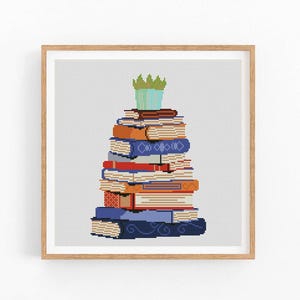 May include: A framed cross-stitch design featuring a stack of colorful books. The books are in various colors, including blue, orange, and red. A small green and blue plant sits atop the stack. The artwork is set against a white background.