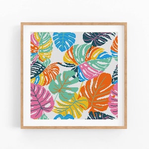May include: A framed artwork featuring a colorful pattern of monstera leaves. The leaves are rendered in a cross-stitch style, with vibrant hues of pink, orange, yellow, blue, and green. The artwork is set against a white background and framed in a light brown wooden frame.