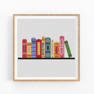 May include: A framed print showing a row of colourful, stylised books on a black shelf. The books have different designs and colours, with one labelled "DETECTIVE" and another "LOVE STORY". The background is a light grey grid.