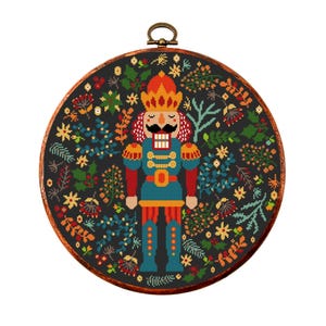 May include: A cross-stitch pattern featuring a colourful nutcracker standing in the centre of a wreath of flowers and leaves. The nutcracker is wearing a blue and red uniform with gold accents. The background is black.