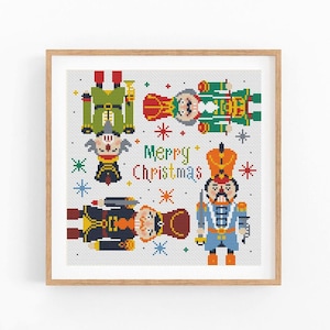 Può includere: Un motivo a punto croce quadrato in una cornice di legno chiaro che raffigura tre schiaccianoci in uniformi verdi, blu e blu scuro. Il testo Merry Christmas è centrato in rosso e verde, circondato da piccoli accenti a forma di stella.
