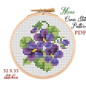 May include: A cross stitch pattern featuring a cluster of purple and green violets. The pattern is 32 stitches wide by 33 stitches high. The pattern is displayed in a wooden embroidery hoop.