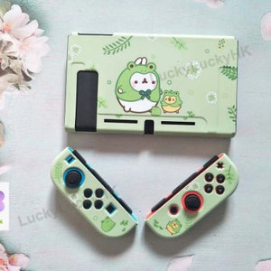 Puede incluir: Un estuche y un conjunto de mandos para consola Nintendo Switch de color verde claro. El estuche y los mandos presentan un dise&ntilde;o de rana de dibujos animados con detalles florales blancos. Los mandos tienen detalles azules y rojos.