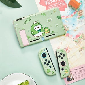 Puede incluir: Una funda y mandos de Nintendo Switch de color verde claro con un dise&ntilde;o de dibujos animados de rana. La funda y los mandos tienen un dise&ntilde;o a juego con ilustraciones de ranas y flores. Los mandos tienen botones y joysticks negros.